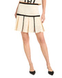 Waist down image of a person wearing a cream pleated skirt with black zigzag stitching and black pointed-toe heel