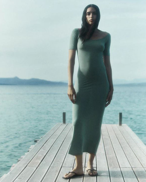 A brunette model standing on a pier next to the water wearing a sage green short-sleeve dress with a pair of black flip flops.