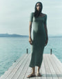 A brunette model standing on a pier next to the water wearing a sage green short-sleeve dress with a pair of black flip flops.