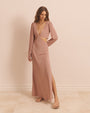 A full body angled view of a model with her hand behind her back, wearing a long-sleeved dress with two triangular cut-out details at the waist and a low V-neck. The model is wearing brown strap up sandals, and long necklaces. The background is plain pink.