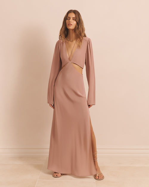 A full body view of a model wearing a long-sleeved dress with two triangular cut-out details at the waist and a low V-neck. The model is wearing brown strap up sandals, and long necklaces. The background is plain pink.