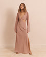 A full body view of a model wearing a long-sleeved dress with two triangular cut-out details at the waist and a low V-neck. The model is wearing brown strap up sandals, and long necklaces. The background is plain pink.