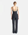 A backside view of a blonde-haired model wearing a bun, a black sleeveless shirt featuring thin straps with an open back, and an opening with a strap crossing at the back. The shirt is paired with black pants, while standing against a white background.