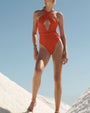 A body shot of a model in front of a blue-sky background, standing on sand dunes wearing a red one-piece swimsuit with a halter neck design, a cut-out diamond detail at the center, and horizontal gathered fabric across the midsection. The background is plain white.