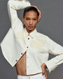 A brunette model with her hand on her head and one on her hip wearing a collared white jacket with doily/floral embroidery on the upper chest. The background is plain gray.