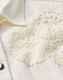 A close-up view of a white collared shirt with a silver button and a crocheted floral lace pattern on the chest area.