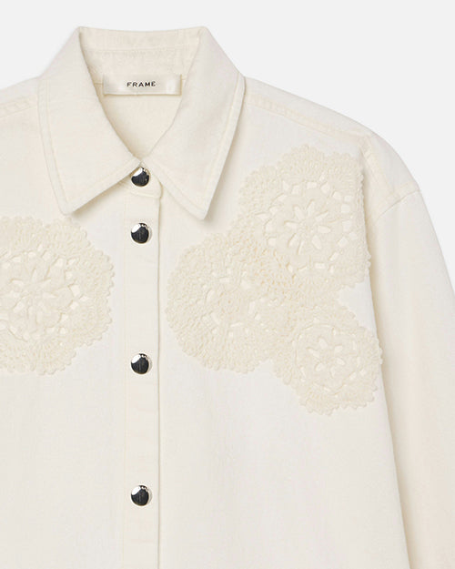 A close image of a collared white jacket with doily/floral embroidery on the left and right side of the upper chest. The jacket has silver buttons and a tag in the inner collar reading "FRAME".