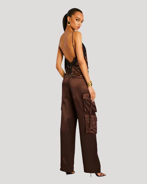 A full body view of a brunette model with her ponytail swept to the side, wearing large gold earrings, three gold bangles, and a thin strapped V-neck sequin top with lace detailing at the top. The model is wearing brown trousers to match and standing in front of a white background.