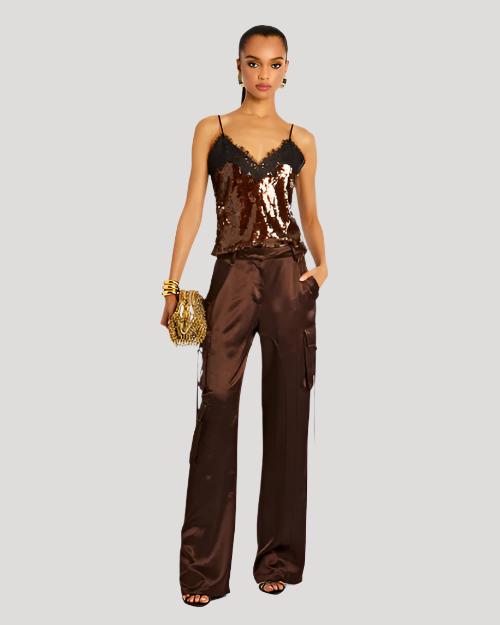 A full body view of a brunette model wearing large gold earrings, three gold bangles, a gold purse and a thin strapped V-neck sequin top with lace detailing at the top. The model is wearing brown trousers to match and standing in front of a white background.
