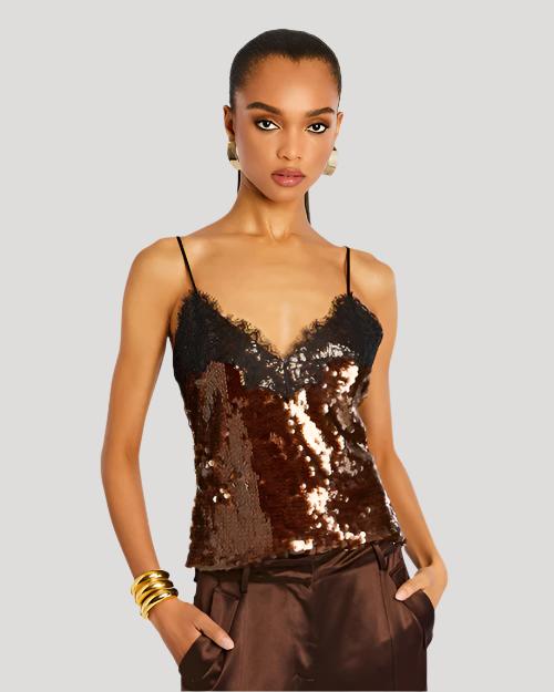 A waist-up view of a brunette model wearing large gold earrings, three gold bangles, and a thin strapped V-neck sequin top with lace detailing at the top. The model is wearing brown trousers to match and standing in front of a white background.