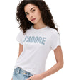 A curly-haired brunette model against a white background wearing a white t-shirt with the text "J'ADORE" written on it in large, light blue letters, paired with light blue jeans.