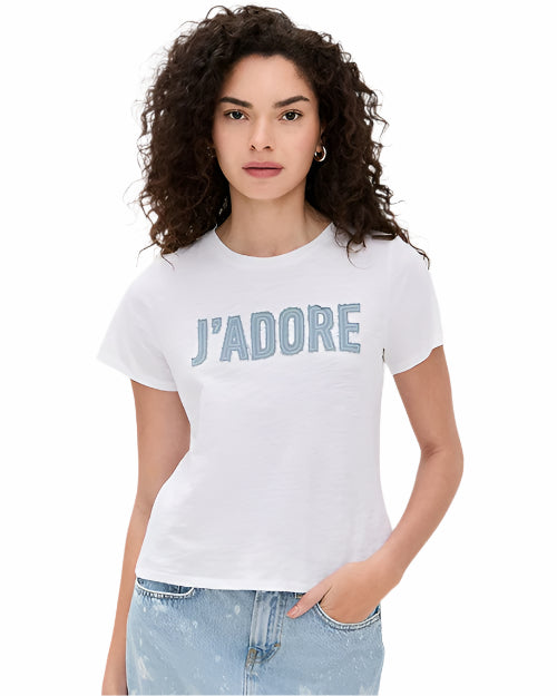 A curly-haired brunette model against a white background, wearing a white t-shirt with the text "J'ADORE" written on it in large, light blue letters, paired with light-blue jeans.