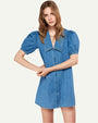 A brunette model is wearing a blue denim dress with short, puffed sleeves, a large, pointed collar and denim buttons down the front, standing in front of a white background.