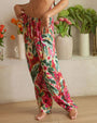 A legs-only view of a model with her hands on her hips wearing loose-fitting pants with an elastic waistband and drawstrings, and a watercolor floral pattern. The model is standing barefoot on a wooden floor, in a homely setting with multiple indoor plants behind her.