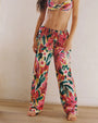 A body only shot of a model wearing a matching watercolor floral pattern set consisting of colorful bikini top with a fabric rose detail in the center, and loose-fitting pants with an elastic waistband and drawstrings. The model is barefoot and standing in front of a white background.