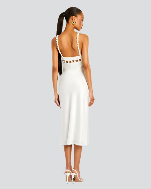 A full body backside view of a brunette model with a ponytail wearing a white midi dress with an open back, thin straps lined with pearls, and cut-out square details along the upper waistline. The model is also wearing white heels, and gold hoops, standing in front of a white background.