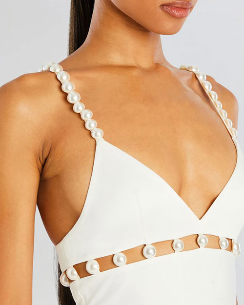 A close-up chest level view of a brunette model wearing a white midi dress with thin straps lined with pearls and cut-out square details with pearls along the upper waistline. The background is plain white.