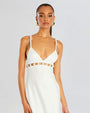 A waist-up view of a brunette model with a ponytail wearing a white midi dress with thin straps lined with pearls and cut-out square details with pearls along the upper waistline. The model is also wearing gold hoops and standing in front of a white background.