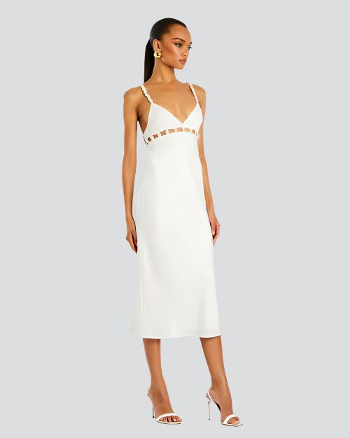 A full body view of a brunette model with a ponytail wearing a white midi dress with thin straps lined with pearls and cut-out square details along the upper waistline. The model is also wearing white open-toed heels, and gold hoops, standing in front of a white background.