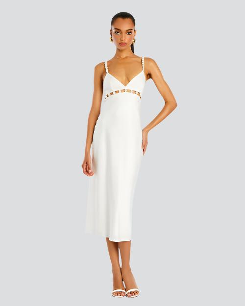 A full body view of a brunette model with her hand on her hip wearing a white midi dress with thin straps lined with pearls and cut-out square details along the upper waistline. The model is also wearing white open-toed heels, and gold hoops, standing in front of a white background.