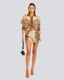 A full body frontal view of a blonde model wearing a matching button-down shirt and shorts with a polka-dot and foliage design, paired with beige open-toed heels and a brown purse. The background is white.