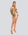 A full body view of a blonde model wearing a matching button-down shirt and shorts with a polka-dot and foliage design, paired with beige open-toed heels. The background is white.