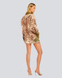 A full body backside view of a blonde model with her hair swept to the side, wearing a matching button-down shirt and shorts with a polka-dot and foliage design, paired with beige open-toed heels. The background is white.