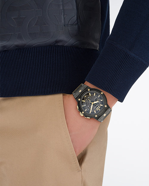 A close-up image of a models' hand in his khaki pocket, wearing a blue leather jacket with an embossed pattern, and a black sporty watch with gold accents. The background is plain white.