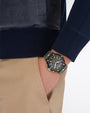 A close-up image of a models' hand in his khaki pocket, wearing a blue leather jacket with an embossed pattern, and a black sporty watch with gold accents. The background is plain white.