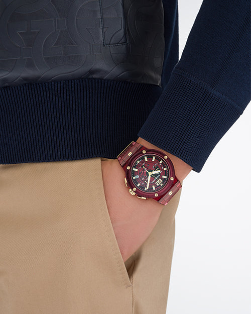 A close-up image of a models' hand in his khaki pocket, wearing a blue leather jacket with an embossed pattern, and a red sporty watch with gold accents. The background is plain white.