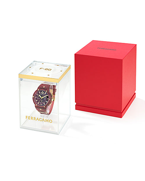 A red sporty wristwatch with gold accents, in clear box with gold detailing and "FERREGAMO" on the front, and "F-80" on the top. The watch is next to a red box with the brand name "FERREGAMO" on the top in black. The background is plain white.