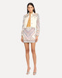 A dark-haired model wearing a yellow shirt under a white denim jacket with intricate, colorful embroidery on the shoulders and sleeves and a matching skirt. The model is wearing matching embroidered skirt with silver open-toed heels.