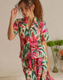 A close photo of a curly-haired brunette model with her hand touching her bangs, wearing a matching watercolor floral patterned button-down short-sleeve shirt, and flowy pants. The model is standing in front of a white wall with indoor plants.