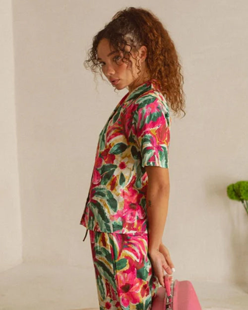 A knee-up sideview of a curly-haired brunette model with her hands behind her back, holding a pink briefcase and wearing a matching watercolor floral patterned button-down short-sleeve shirt, and flowy pants. The model is standing in front of a white wall with a green indoor plant.