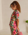 A knee-up sideview of a curly-haired brunette model with her hands behind her back, holding a pink briefcase and wearing a matching watercolor floral patterned button-down short-sleeve shirt, and flowy pants. The model is standing in front of a white wall with a green indoor plant.
