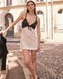 A model in front of a white building and holding onto a black railing is wearing a V-neck dress with thin black straps, black lace detailing, a bow on the upper half, and a white fabric lower section. The background is plain white.