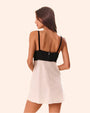 A backside knee-up view of a brunette model with her hair swept to the side, wearing a V-neck dress with thin black straps, a half open back, and a white fabric lower section. The background is plain white.