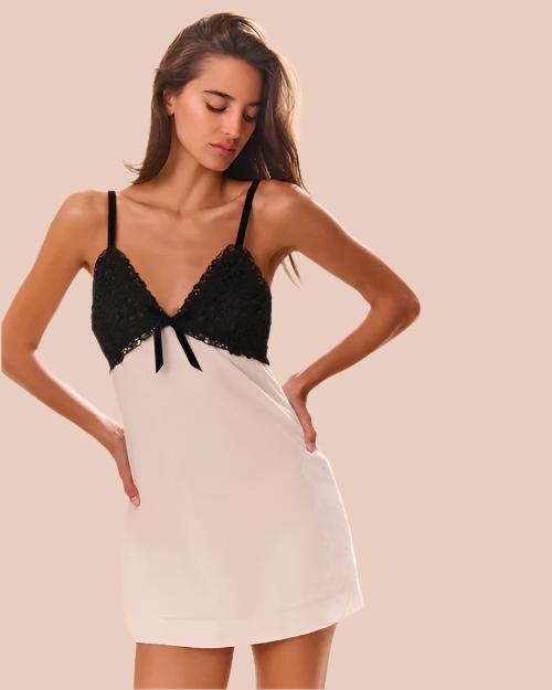 A knee-up view of a model with her hands on her hips, wearing a V-neck dress with thin black straps, black lace detailing with a bow on the upper half, and a white fabric lower section.  The background is plain white.