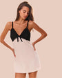 A knee-up view of a model with her hands on her hips, wearing a V-neck dress with thin black straps, black lace detailing with a bow on the upper half, and a white fabric lower section.  The background is plain white.
