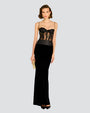 A short-haired brunette wearing a long dress with a black lace corset top, thin straps and intricate lace detailing around the waist. The lower half is a velvet black skirt, and the model accessorized with a gold clutch and black heels. The model paired the dress with gold earrings, a gold square clutch, and black heels while standing against a white background.