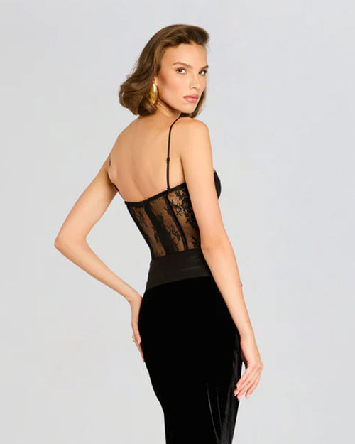 An angled backside view of a short-haired brunette model wearing gold earrings, a black dress with a lace bodice featuring intricate detailing, with thin straps and a low cut back. The lower half is a velvet black skirt, and the background is plain white.