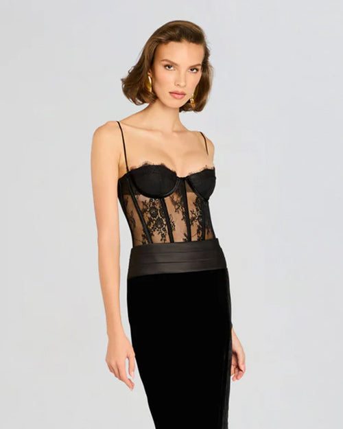 A short-haired brunette model wearing gold earrings and a dress with a black lace corset top, thin straps and intricate lace detailing around the waist. The lower half is a velvet black skirt, and the background is plain white.