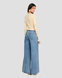 The backside view of a dark-haired model wearing a cream, long-sleeved crochet cardigan with intricate geometric patterns, paired with blue jeans, heels, and large gold earrings, while the model is standing against a white background.