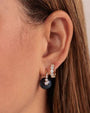 A close-up view of a brunette models' ear wearing a dark blue spherical gemstone earring, attached to a gold hoop adorned with a linear arrangement of round diamonds. Next to it is a gold hoop adorned with white diamonds.