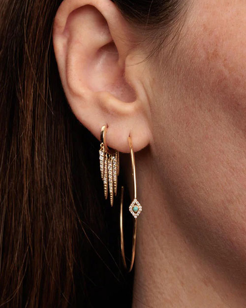 A close-up image of a brunette models' earring wearing a gold hoop earring with jewel adorned spikes hanging from the hoops, and thinner gold hoop under it decorated with a diamond jewel design in the center.
