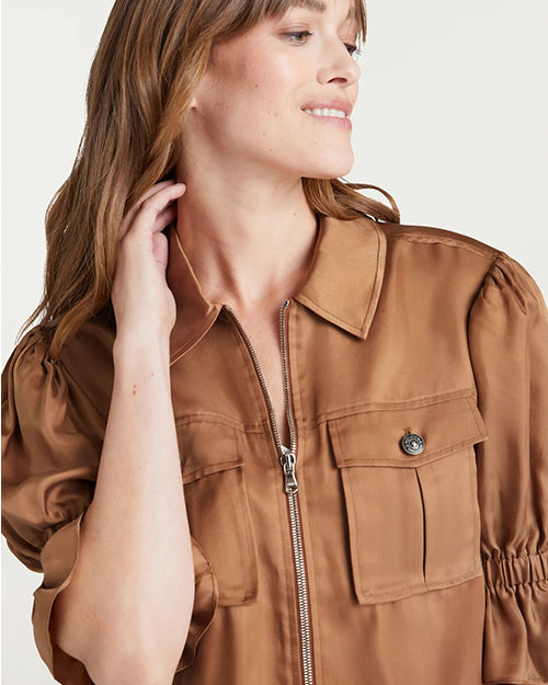 A waist-up image of a brunette model with her hand to her neck, wearing a brown jacket with a zipper and chest pockets, and bunching at the elbows. The background is plain white.