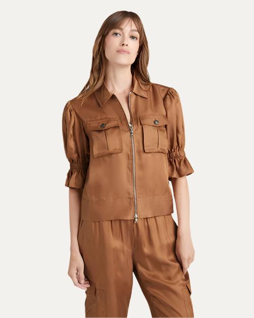 A knee-up image of a brunette model wearing a brown jacket with a zipper, chest pockets, and bunching at the elbows. The model is wearing matching brown pants and standing in front of a white background.