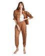 A full body image of a brunette model wearing an opened brown short-sleeve jacket with a zipper, chest pockets, and bunching at the elbows. The model is wearing a white shirt under and matching brown elastic banded pants while standing in front of a white background.