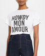 A torso level shot of a model wearing a white crewneck t-shirt with the text "HOWDY MON AMOUR" printed on it in black capital letters, adorned with small gray dots. The model is also wearing high-waisted dark blue jeans and gold hoop earrings, while standing in front of a white background.