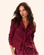 A waist-up view of a brunette model wearing a long-sleeved, button-up, maroon blouse with intricate floral and geometric patterns. The shirt is paired with matching bottoms, and the model is standing in front of a white background.